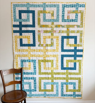 Modern quilt with looping geometric shapes in blue, green, and yellow tones hanging on a wall beside a wooden chair, showcasing the Line Dance Quilt Pattern (Download) by Penny Spool Quilts.