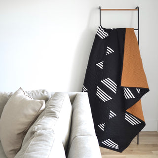 Living Room Quilt Pattern (Download) by Sewn displayed on a modern black ladder with geometric black and white design and warm brown backing in a cozy living room setting.