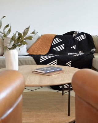 Cozy living room scene featuring a black and white geometric quilt draped over a beige sofa, a round wooden coffee table with books, and a white vase with green leaves, styled by Sewn using the Living Room Quilt Pattern (Download).