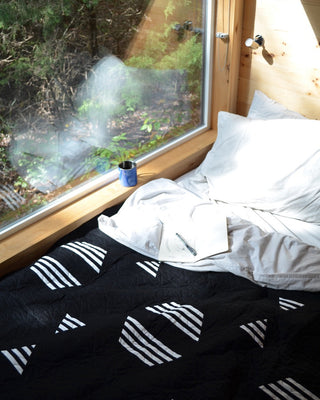 Cozy bedroom scene featuring a black and white Living Room Quilt Pattern (Download) by Sewn on the bed near a large window with natural light and a blue mug.