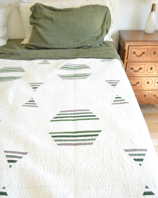 Bright bedroom with a white quilt featuring geometric green and gray striped shapes, styled with olive green pillows and a wooden nightstand, showcasing the Living Room Quilt Pattern (Download) by Sewn.