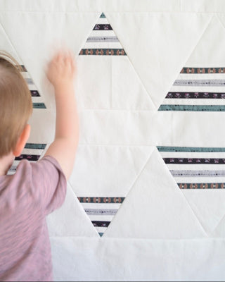 A young child reaching toward a white quilt featuring triangular shapes with patterned fabric strips, showcasing the Living Room Quilt Pattern (Download) by Sewn.