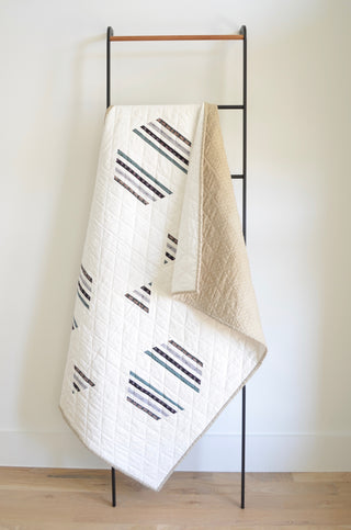 Light-colored quilt with geometric stripe block patterns hanging on a modern black ladder, showcasing the Living Room Quilt Pattern (Download) by Sewn.