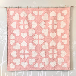Pink and white heart-themed quilt hung on a wall, showcasing intricate stitching in a modern geometric design, made with The Dandy Patch Lovelace Quilt Pattern (Download).