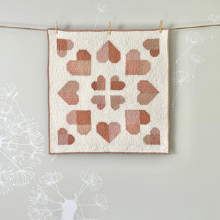 Soft pink and beige heart-shaped quilt blocks arranged in a symmetrical pattern on a white background hanging with clothespins, showcasing the Lovelace Quilt Pattern (Download) by The Dandy Patch.