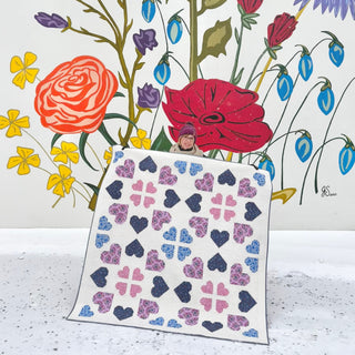 Person holding a colorful quilt with heart and clover shapes in a snowy outdoor scene against a vibrant floral mural, showcasing The Dandy Patch Lovelace Quilt Pattern (Download).