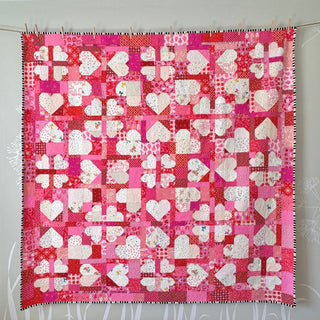 Bright pink and red scrappy quilt featuring white heart and plus block patterns, showcasing a vibrant handmade design by The Dandy Patch, created with the Lovelace Quilt Pattern (Download).