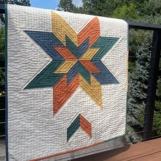 Max Patch Quilt Pattern by Hampton Quilt Studio hanging: bold colorway with orange, teal, and gold geometric star on white background, displayed on a deck railing outdoors.