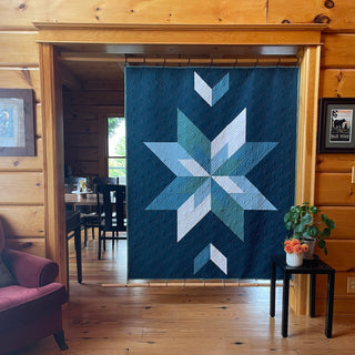 Max Patch Quilt Pattern by Hampton Quilt Studio lifestyle: teal and navy geometric star quilt hanging in a log cabin room with wood walls, hardwood floors, and a plant.