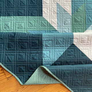 Max Patch Quilt Pattern by Hampton Quilt Studio closeup: geometric star quilt in teal, navy, and white on wood surface, corner folded to show mint green backing.