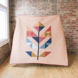 A modern quilt with geometric leaf shapes in muted colors displayed against a brick wall, showcasing the May Quilt Pattern (Download) by Month of May Quilting.