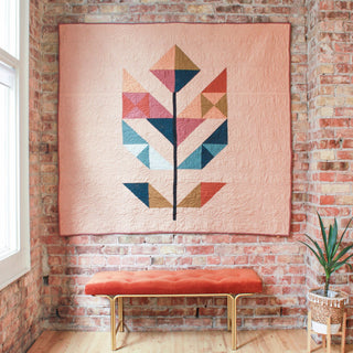 Modern quilt with geometric leaf design in soft pink, blue, mustard, and rust tones hanging on a brick wall above an orange bench, showcasing the May Quilt Pattern (Download) by Month of May Quilting.