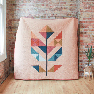 Cozy quilt with geometric flower pattern in warm and cool tones against a soft pink background, showcasing the May Quilt Pattern (Download) by Month of May Quilting.