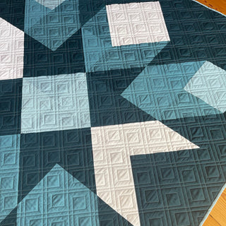 Mt. Mitchell Quilt Pattern by Hampton Quilt Studio flat lay: overhead closeup of navy, teal, and white geometric diamond star quilt on hardwood floor showing quilting detail.