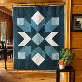 A modern geometric quilt in shades of blue and white hanging in a cozy wooden room, showcasing the Mt. Mitchell Quilt Pattern (Download) by Hampton Quilt Studio.