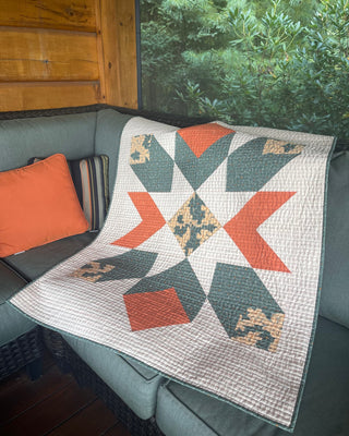 Quilt featuring a geometric star pattern in muted orange, green, and beige draped on a porch sofa with cushions, showcasing the Mt. Mitchell Quilt Pattern (Download) by Hampton Quilt Studio.