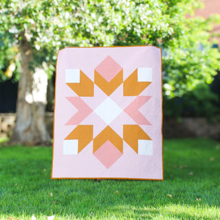 Mt. Mitchell Quilt Pattern (Download) by Hampton Quilt Studio featuring a pink and golden geometric quilt with half square triangles displayed outdoors