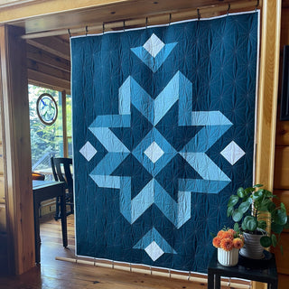Large geometric quilt with a star pattern in shades of blue and white, hanging in a cozy wooden room, showcasing the Mt. Pisgah Quilt Pattern (Download) by Hampton Quilt Studio.