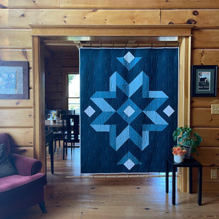 Hanging quilt featuring a bold geometric star pattern in shades of blue, displayed in a cozy wooden cabin setting by Hampton Quilt Studio using Mt. Pisgah Quilt Pattern (Download).