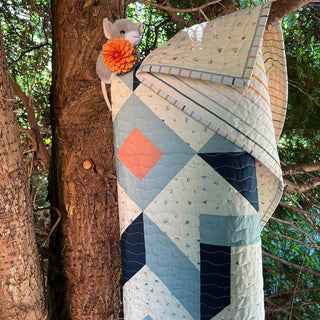 Quilt draped on tree featuring soft blue, coral, and navy geometric patterns with a plush mouse toy and orange flower, showcasing the Mt. Pisgah Quilt Pattern (Download) by Hampton Quilt Studio.