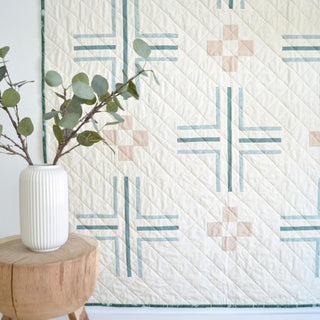 Mudroom Quilt Pattern by Sewn blue colorway: cream quilt with teal and blush cross and pinwheel blocks hung on a wall, styled with a white vase of eucalyptus.