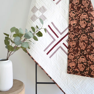 Close-up of a modern geometric quilt with muted tones draped on a black ladder rack next to a white vase with eucalyptus, showcasing the Mudroom Quilt Pattern (Download) by Sewn.