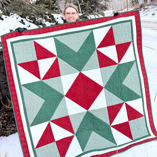 A person outdoors in winter holds a red, green, and white quilt showcasing geometric star and triangle patterns, featuring the Northern Glow Quilt Pattern (Download) by Shadow Valley Quilts.
