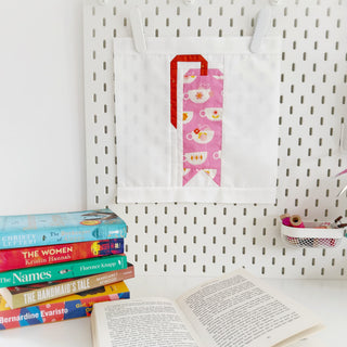 White fabric quilt block with a pink teacup and red bookmark design hanging on a pegboard, part of One More Page Block Pattern (Download) by Lou Orth Designs.