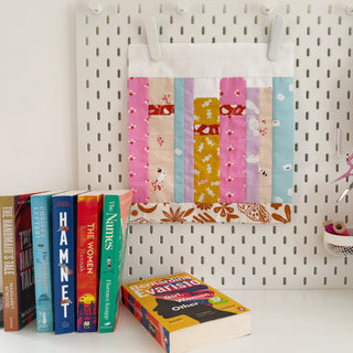 Colorful fabric quilt block with patterns inspired by books on a pegboard, showcasing On the Shelf Block Pattern (Download) by Lou Orth Designs in a cozy craft space.