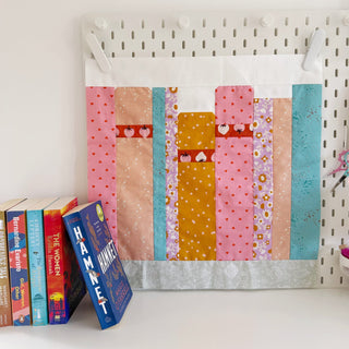 Colorful quilt block with vertical rectangular patterns in pink, mustard, teal, and floral fabrics displayed on a pegboard, featuring "The Book Block Collection — Complete 5-Block Bundle (Download)" by Lou Orth Designs.