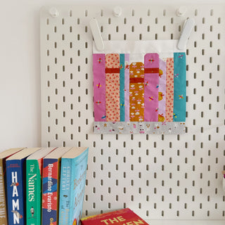 Brightly colored quilt block featuring vertical stripes in pink, orange, blue, and white hangs on white pegboard above stacked books, showcasing On the Shelf Block Pattern (Download) by Lou Orth Designs.