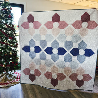 Quilt featuring flower-inspired geometric shapes in muted pinks, blues, and purples displayed next to a decorated Christmas tree, showcasing the Orchid Garden Quilt Pattern (Download) by A Piece of Quiet Quilts.