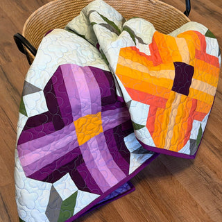 Two colorful quilts featuring large floral patterns in purple and orange tones folded in a basket, showcasing the Pansy Picnic Quilt Pattern (Download) by Shadow Valley Quilts.