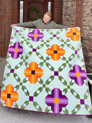 Person holding a colorful quilt with orange and purple pansy-inspired blocks connected by green geometric shapes, showcasing the Pansy Picnic Quilt Pattern (Download) by Shadow Valley Quilts.