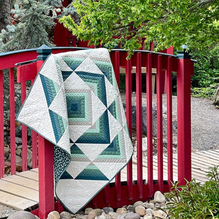 A quilt with geometric patterns in shades of green, blue, and white draped over a red wooden bridge outdoors, showcasing the Percussion Quilt Pattern (Download) by Shadow Valley Quilts.