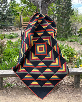 Colorful geometric quilt with concentric square patterns in warm tones draped outdoors on a bench, showcasing the Percussion Quilt Pattern (Download) by Shadow Valley Quilts.