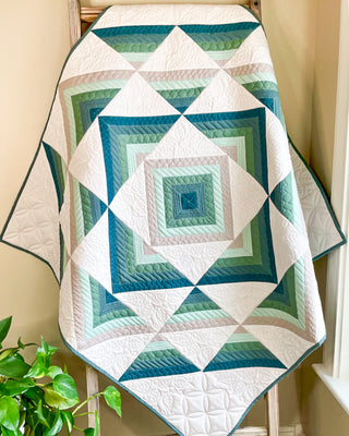 Quilt with geometric green, blue, and beige pattern draped on wooden ladder showcasing Percussion Quilt Pattern (Download) by Shadow Valley Quilts.