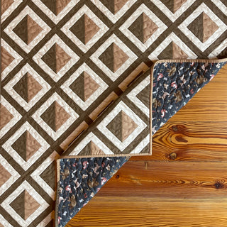 Pine Shadow Quilt Pattern by Hampton Quilt Studio backing: brown and cream diamond shadow quilt on wood surface, corner folded to reveal dark floral backing fabric.