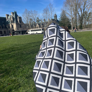 Person holding a gray and white geometric quilt outdoors near a historic stone building, showcasing the Pine Shadow Quilt Pattern (Download) by Hampton Quilt Studio.