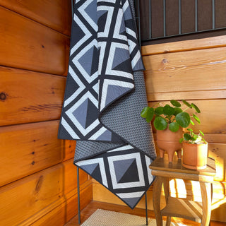 Black, white, and gray geometric quilt draped on a metal stand beside wood-paneled walls with two small potted plants, showcasing the Pine Shadow Quilt Pattern (Download) by Hampton Quilt Studio.
