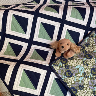 A green, navy, and white geometric quilt made using the Pine Shadow Quilt Pattern (Download) by Hampton Quilt Studio, featuring a floral backing and a small plush dog.