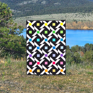 Pivoting Quilt Pattern by Staci W Quilts cover: black and white geometric quilt with rainbow colored squares, displayed outdoors by a mountain lake.