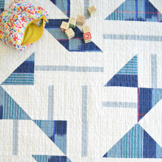 Close-up of a geometric quilt with blue and gray tones, handmade using the Playroom Quilt Pattern (Download) by Sewn, with wooden baby blocks and a colorful fabric pouch.