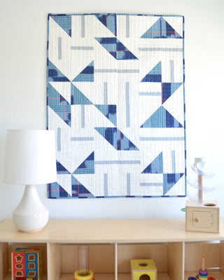 Blue and white geometric quilt with modern flying geese shapes hanging on a wall above a light wood shelf with toys, crafted using Sewn's Playroom Quilt Pattern (Download).