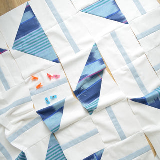 Close-up of a modern quilt in progress featuring blue geometric shapes and light gray strips, with colorful sewing clips, showcasing the Playroom Quilt Pattern (Download) by Sewn.