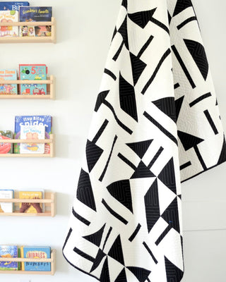Modern black and white quilt draped on a wall, featuring geometric shapes, showcasing the Playroom Quilt Pattern (Download) by Sewn in a bright playroom setting.