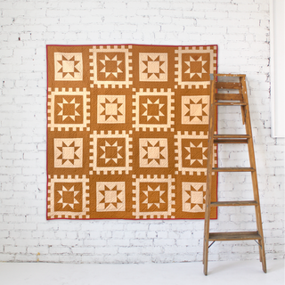 Postmark Quilt Pattern by Sew & Sew Quilting Co. cover: two-tone russet and cream geometric star quilt hanging against a white brick wall with a wooden ladder beside it.