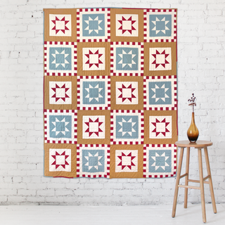 Patchwork quilt with red, blue, and tan geometric star blocks displayed on a white brick wall, styled with a wooden stool and amber vase, showcasing Postmark Quilt Pattern (Download) by Sew & Sew Quilting Co.
