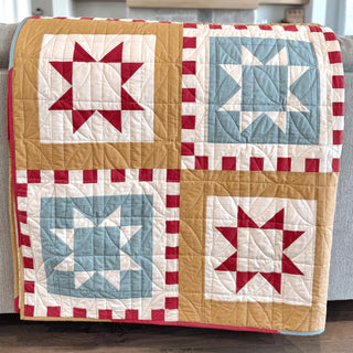 Folded quilt featuring geometric star blocks in red, blue, cream, and mustard colors with a checkerboard border, showcasing the Postmark Quilt Pattern (Download) by Sew & Sew Quilting Co.