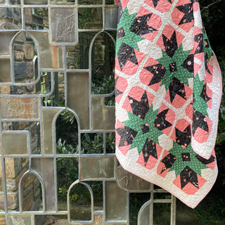 Colorful geometric quilt featuring coral, green, black, and white patterns draped on a decorative glass and stone wall, showcasing the Present Parcels Quilt Pattern (Download) by A Piece of Quiet Quilts.